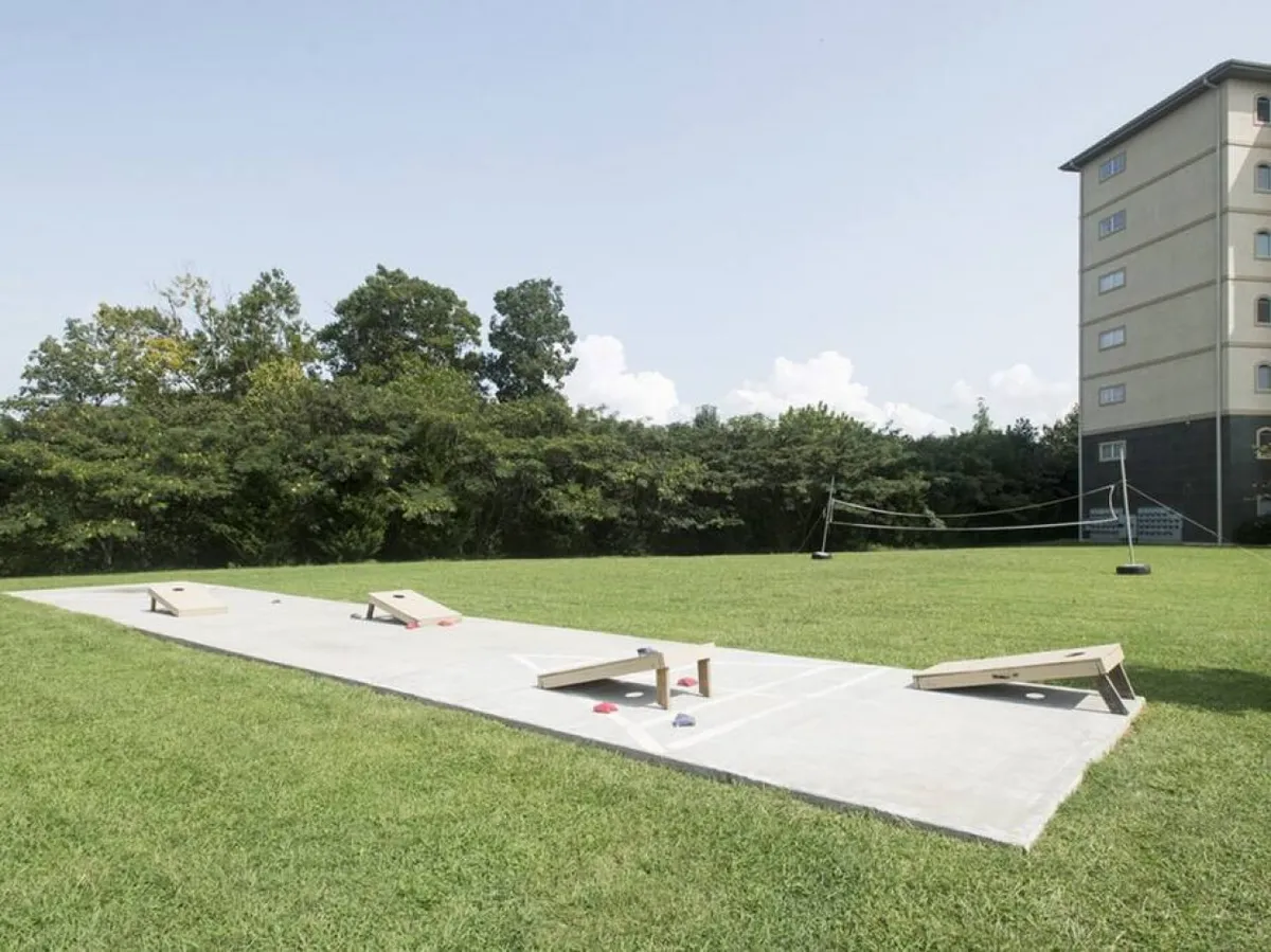 A lively exterior space with cornhole boards set up on lush green grass, a volleyball net in the background, and a tall residential building nearby. The scene is playful, emphasizing group activities and outdoor sports.