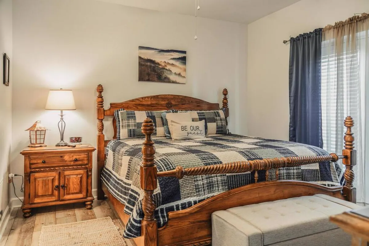 The image showcases a cozy, bear-themed bedroom featuring a plush bed with soft linens, a nightstand with a warm lamp, rustic artwork, and sunlight streaming through thick curtains, creating a tranquil and inviting retreat.