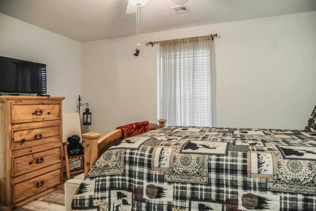 A serene bedroom featuring a bed with a patterned quilt, a wooden dresser, and a window with sheer curtains letting in soft, natural light. The decor is simple yet inviting, ideal for relaxation and rejuvenation.