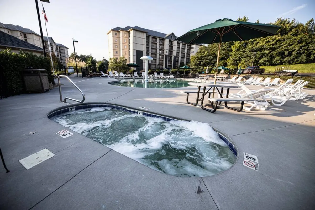 A lively exterior scene showcasing a hot tub, swimming pool, lounge chairs, umbrellas, and a picnic table. The area is set up for relaxation, socializing, and enjoying the outdoors within the property itself.