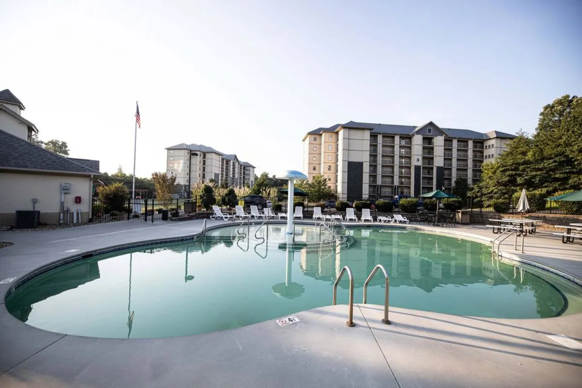 A vibrant outdoor swimming pool scene surrounded by lounge chairs, decorative umbrellas, and lush greenery. The pool area looks inviting for families, with enough space for kids to play and adults to relax. The clear blue water and well-maintained amenities suggest a lively but peaceful place for enjoyment.