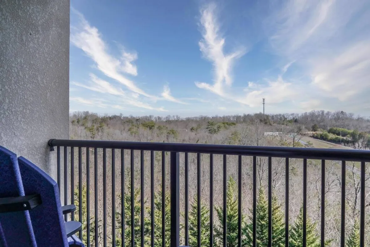 This image displays a tranquil balcony view overlooking lush green trees with a bright blue sky and soft clouds. The scene is peaceful, emphasizing the opportunity to relax outdoors and take in the beautiful natural surroundings from the property itself.