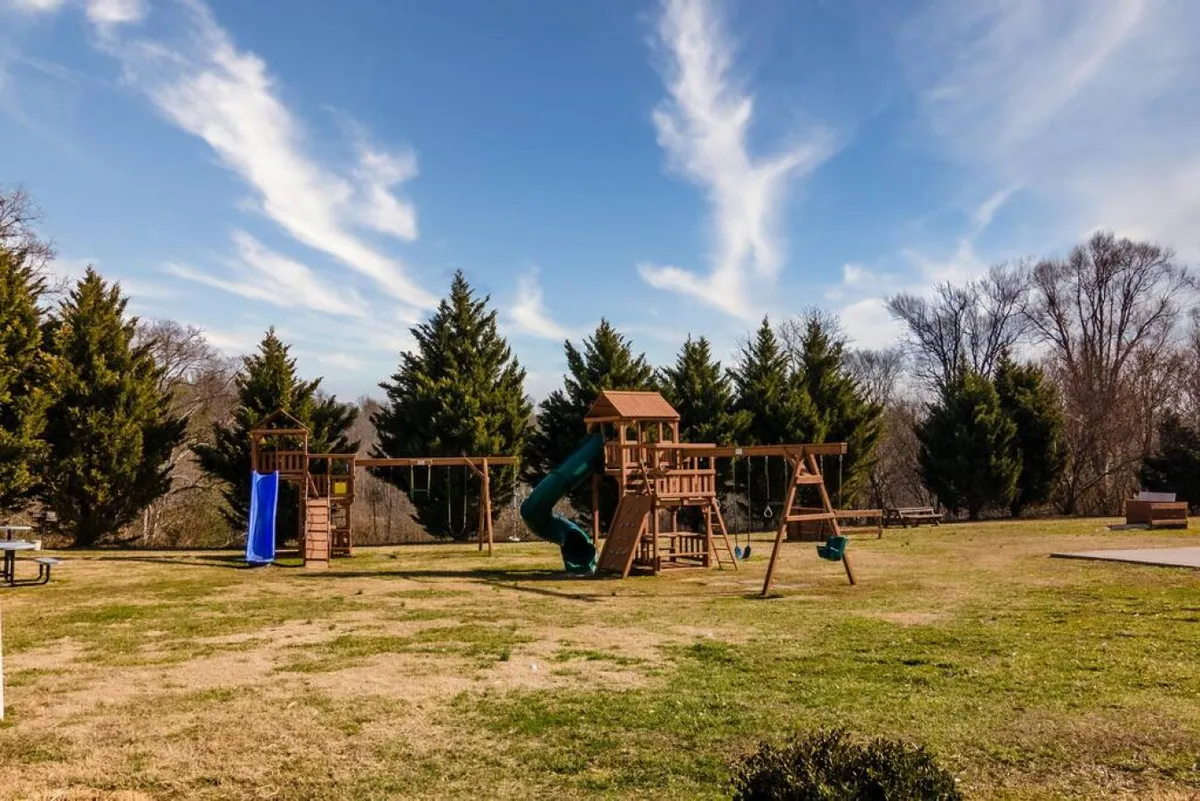 A joyful playground scene with modern equipment including swings, a slide, and an adjacent picnic area on green grass. Families and children are enjoying outdoor games and gatherings in a vibrant, active setting.