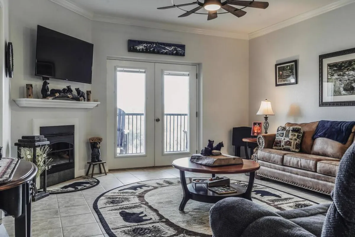 This image features a spacious living room with a comfortable leather sofa, a coffee table, a fireplace for cozy evenings, and double doors leading out to the balcony. The setting is warm and inviting, perfect for gathering, relaxing, or watching TV. The view through the doors hints at the scenic balcony and mountain backdrop.