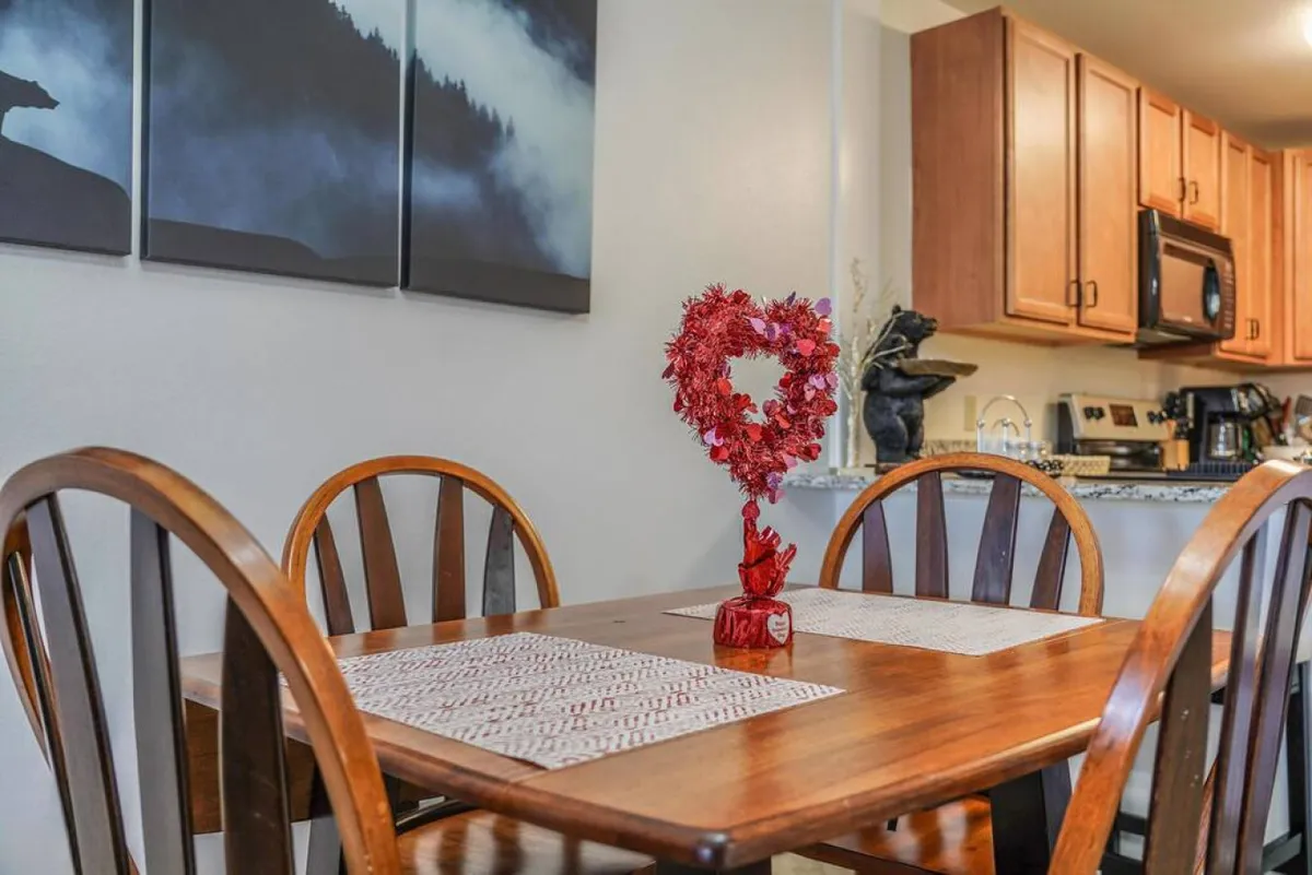 A warm dining space featuring a wooden dining table topped with a heart-shaped floral centerpiece. The kitchen is visible in the background, naturally connecting the dining and cooking areas. The ambiance is cozy, emphasizing togetherness and special occasions.