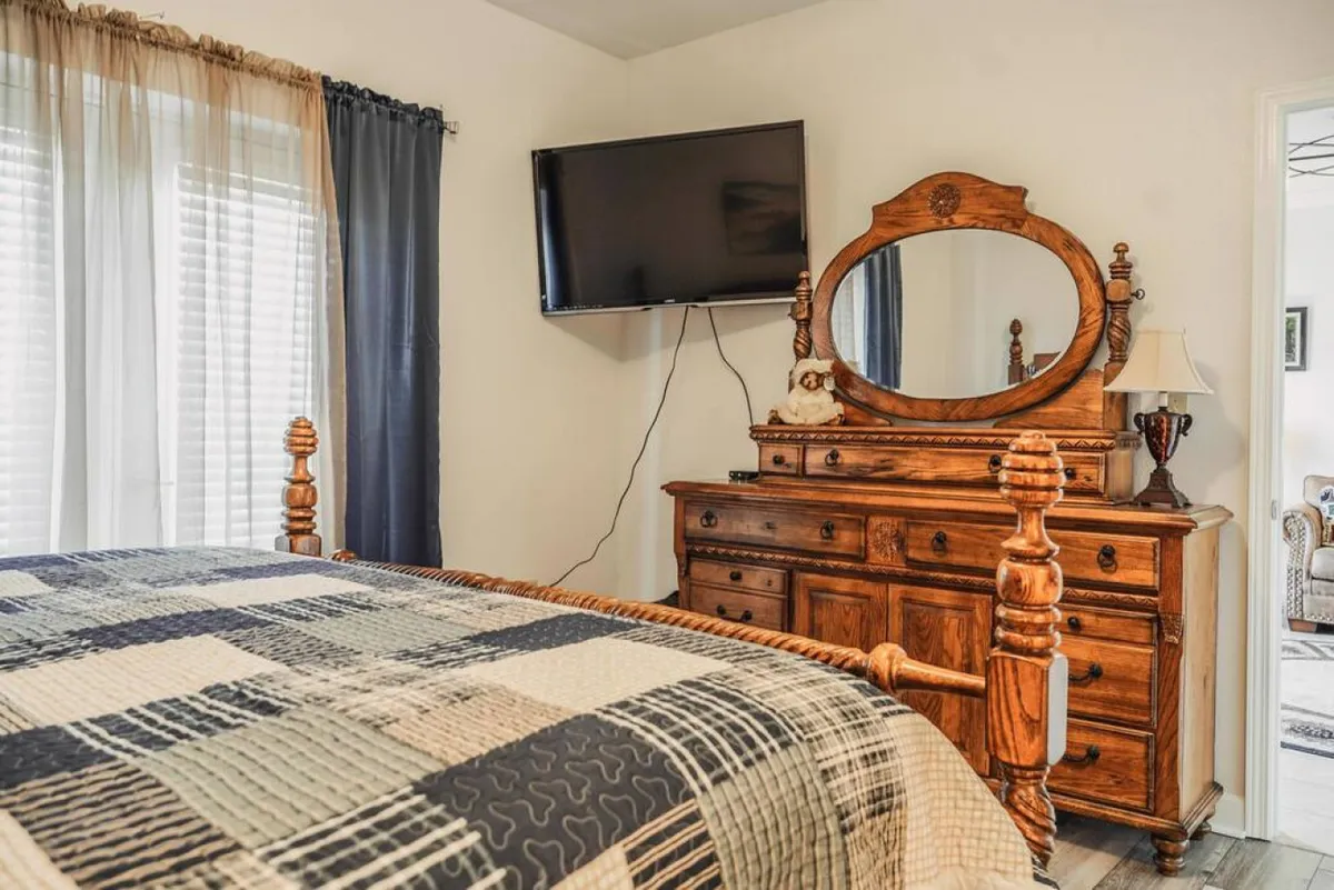 This image showcases a bedroom with a plush bed dressed in a patterned quilt, a wooden dresser with a mirror, and a television mounted on the wall. The soft lighting and warm tones create an inviting and restful atmosphere, ideal for relaxing, watching TV, or enjoying a lazy morning.